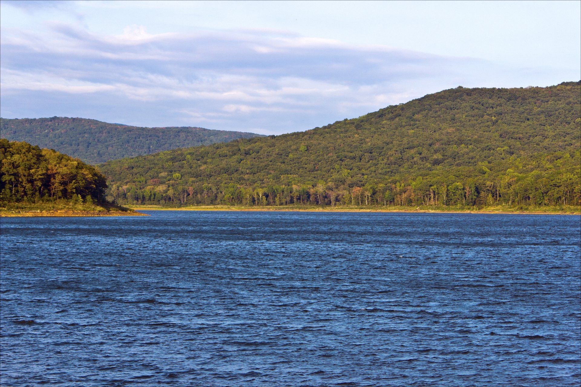 Blue water lake in front of green tree covered mountains under a cloudy sky.