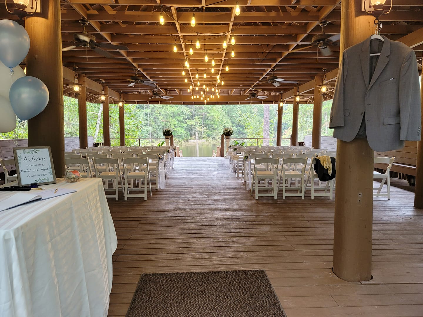 Wedding ceremony setup: white chairs, string lights, gray blazer on a column, overlooking a lake.