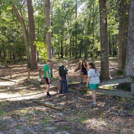 People talking near a weathered wooden fence in a sunny wooded area.