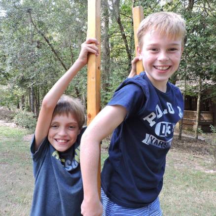Two smiling boys outdoors, holding wooden posts.