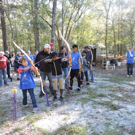 People aiming bows at archery range, wooded area, sunny day.