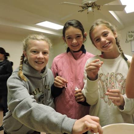 Three girls smiling while doing a science experiment indoors.