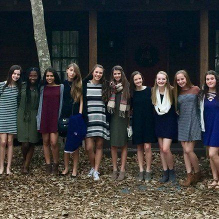 Group of friends smiling in front of a wooden cabin. They are wearing dresses, standing in a line.