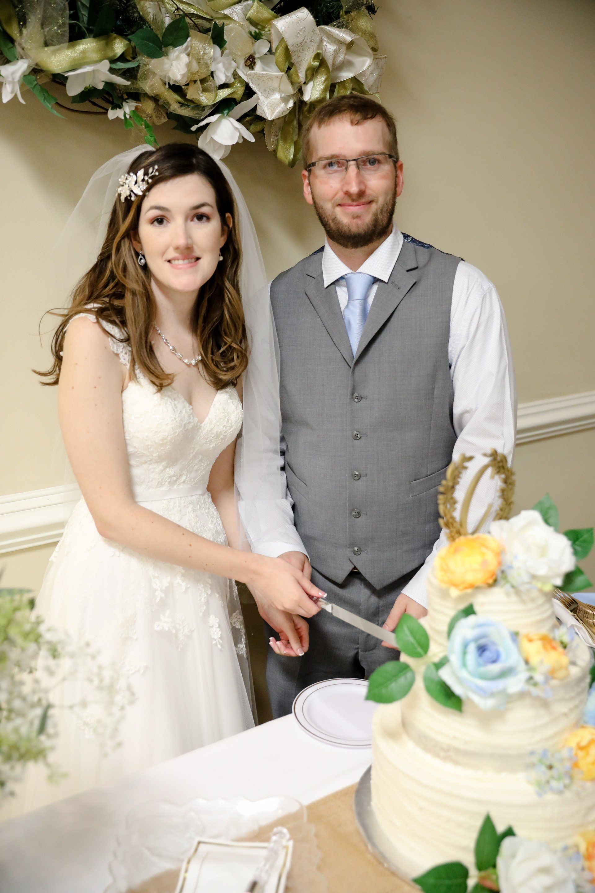 Bride and groom cutting wedding cake. Bride wears white dress, veil, and flower crown. Groom in vest.