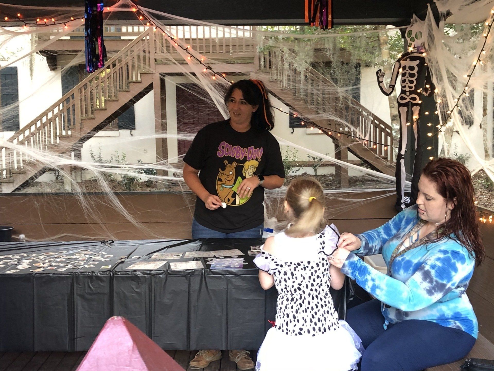 Three people at a Halloween-decorated table. One person hands something to a child, another adjusts the child's costume.
