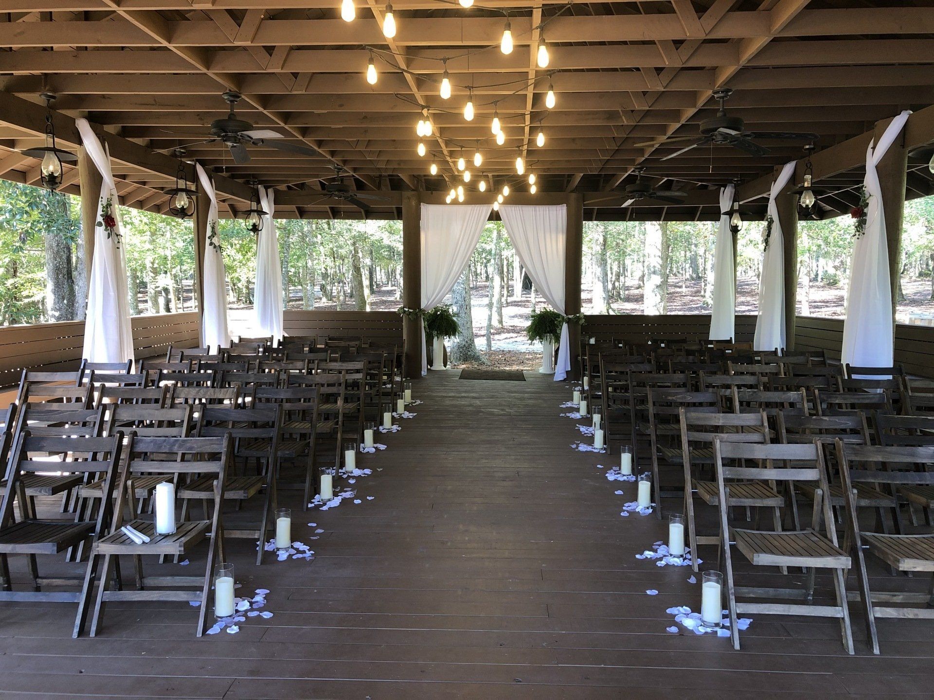 Wedding ceremony setup under a wooden pavilion with rows of chairs, candles, and string lights.
