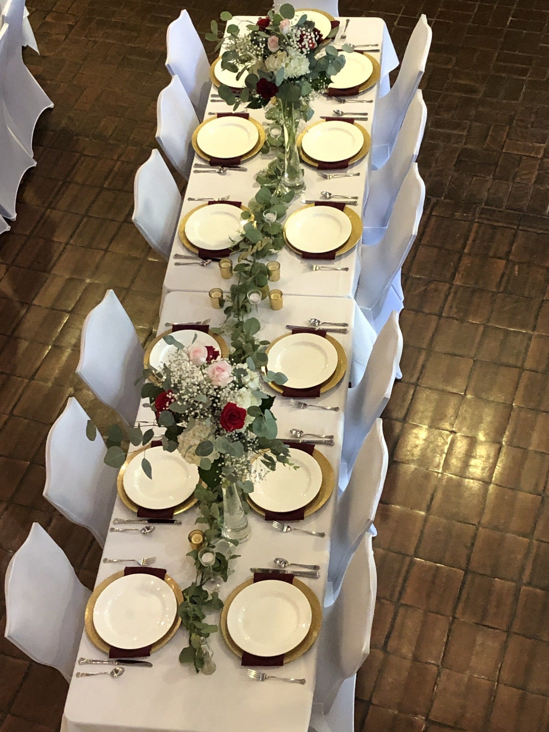 Long rectangular table set for a formal event, decorated with floral arrangements and gold-rimmed plates. White chairs.