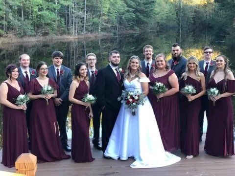 Wedding party posing by a lake; bride and groom centered with bridesmaids in burgundy dresses and groomsmen in grey suits.