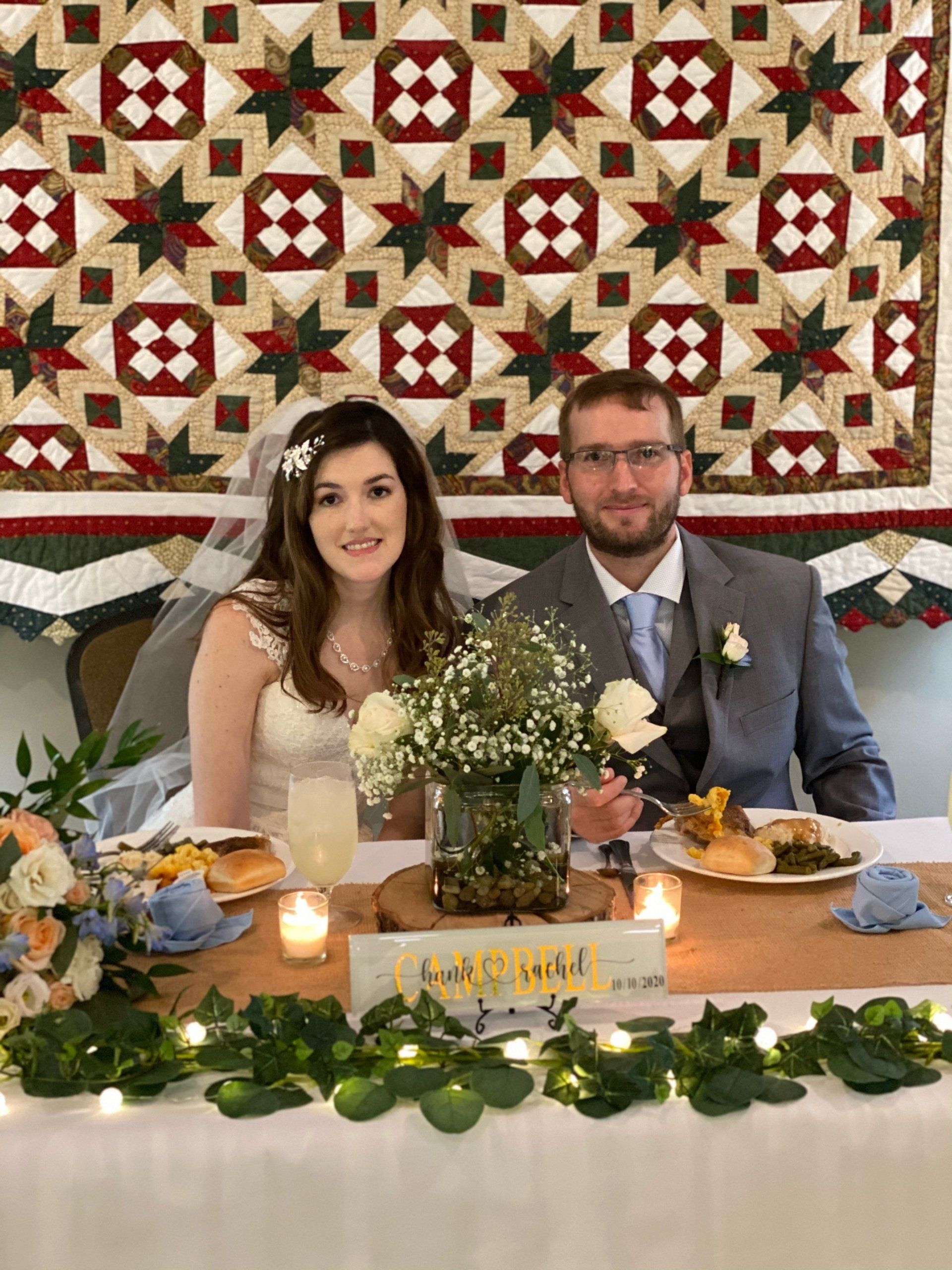 Bride and groom at a wedding reception table; bouquet, candles, and a star-patterned quilt background.