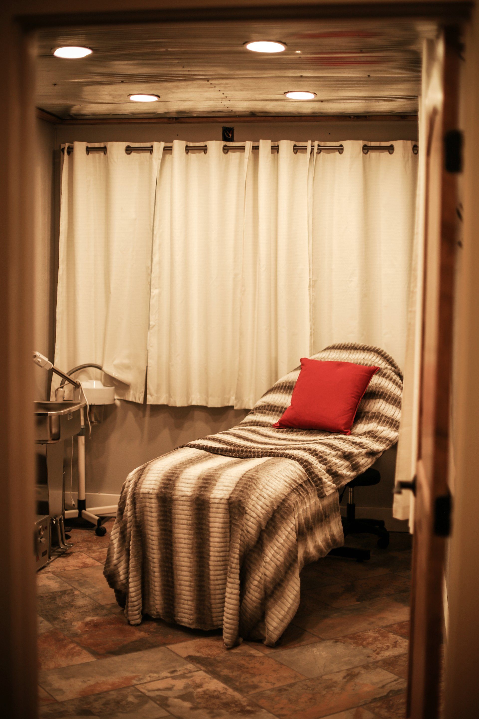Spa treatment room with a reclining chair, red pillow, and cream curtains under warm lighting