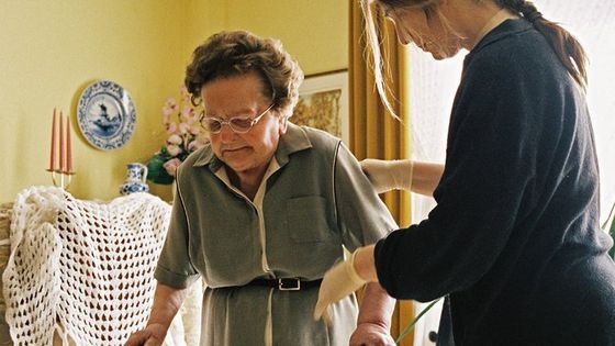 Frau mit Brille wird im Innenbereich von einer Pflegekraft unterstützt.