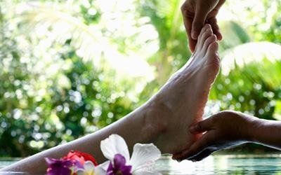 Mani che massaggiano un piede in un ambiente termale con fiori e uno sfondo verde sfocato.
