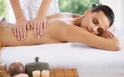 Donna che riceve uno scrub alla schiena in una spa; mani che massaggiano la pelle ricoperta di granuli, candele in primo piano.