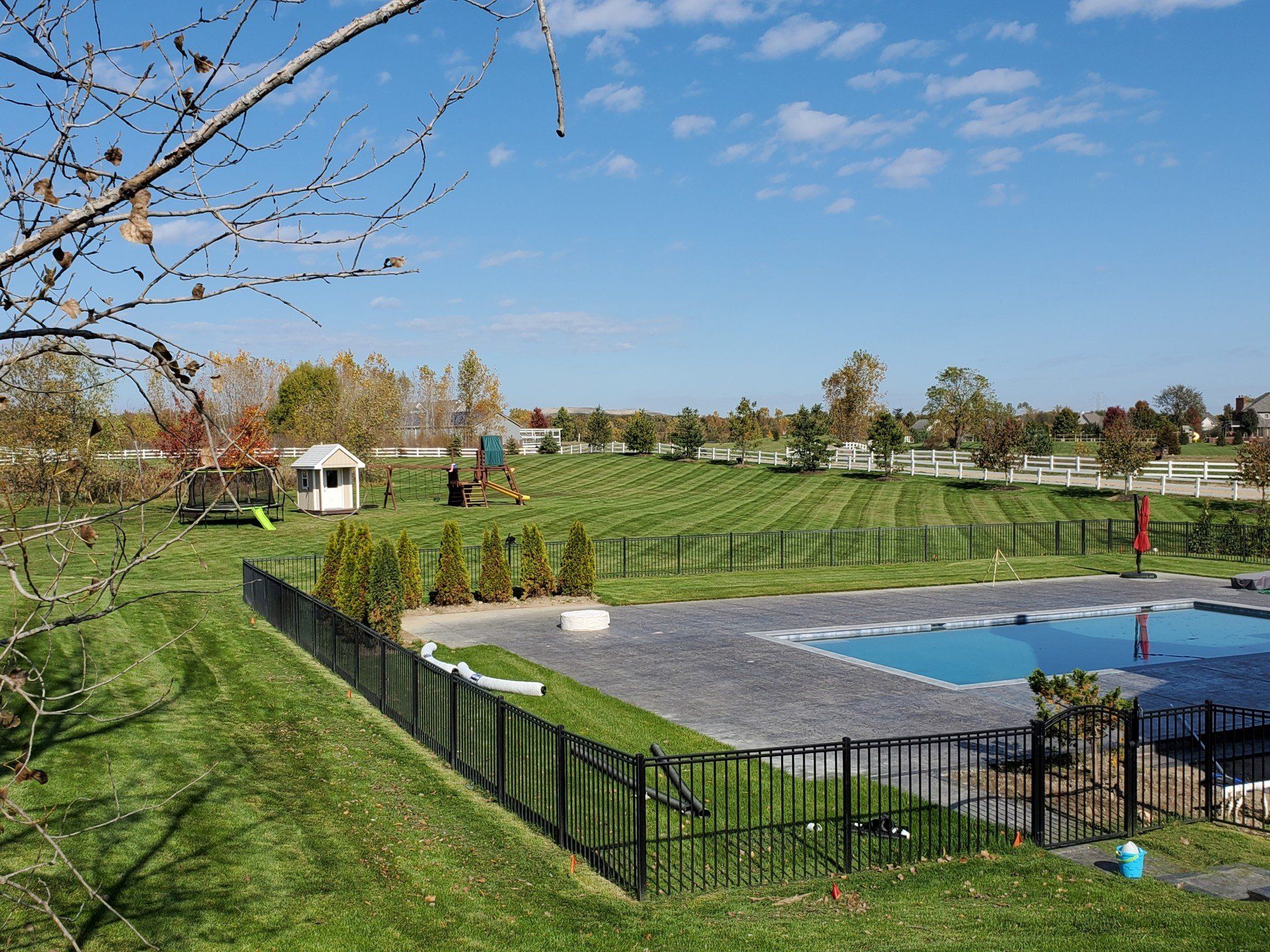 A backyard with a pool, lawn, playground, and trees under a blue sky.