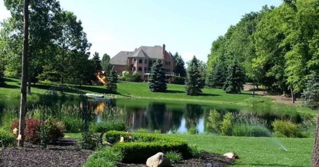 Large house with a pond in front, surrounded by trees and green grass on a sunny day.
