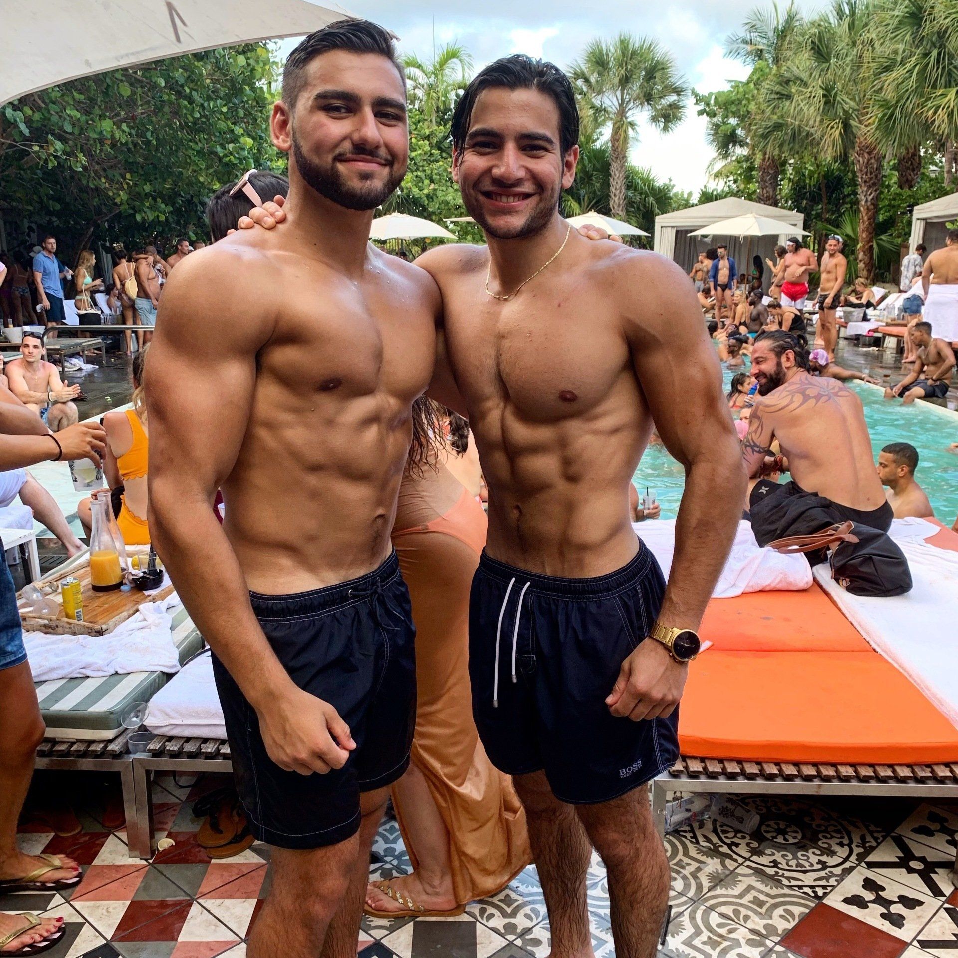 Two shirtless men are standing next to each other in front of a pool.
