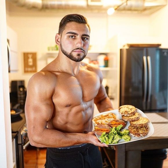 A shirtless man is holding a plate of food in a kitchen.