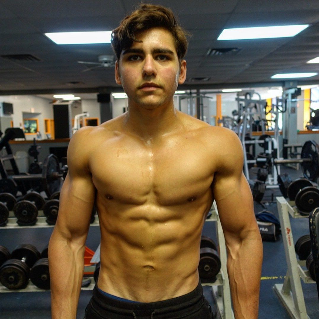 A shirtless man stands in front of a rack of dumbbells in a gym
