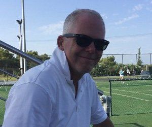 A man wearing sunglasses and a white shirt is sitting on a tennis court.