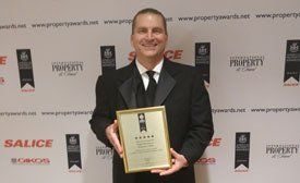 Man in a tuxedo holding an award, smiling in front of a branded backdrop.