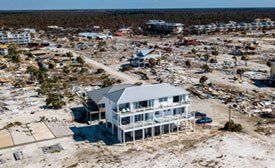 A three-story beach house stands amidst widespread destruction; debris, damaged homes, and barren landscape.