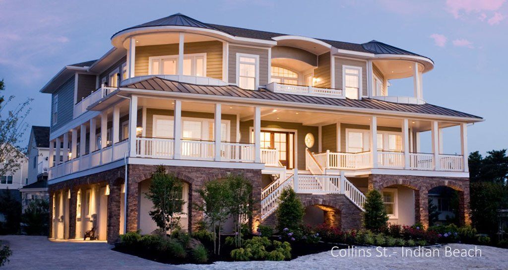 Large, multi-story beach house with ocean view, wrap-around porches, stone base, and blue and tan exterior at dusk.