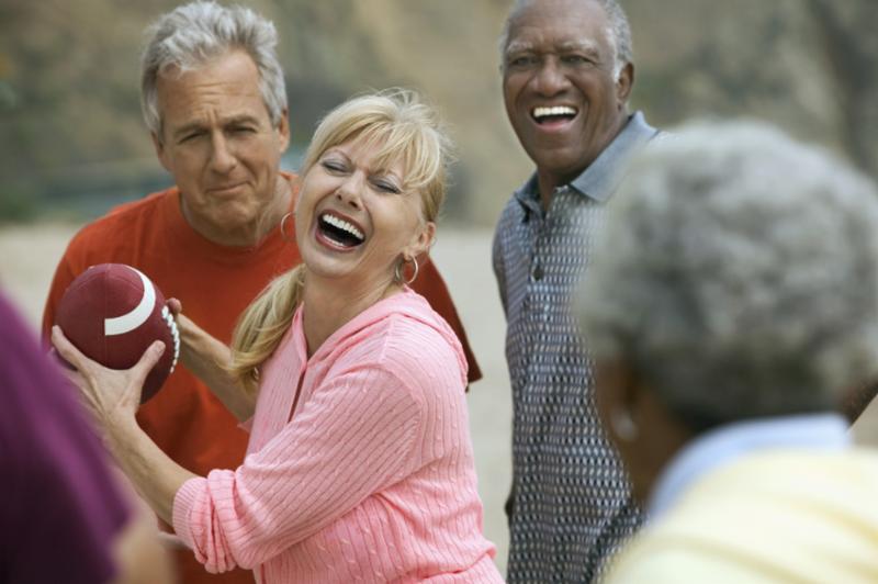 Group of older adults laughing and playing football on a beach, sunny day.