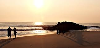 Two people on a beach watch the sunset over the ocean. A rocky breakwater is silhouetted.