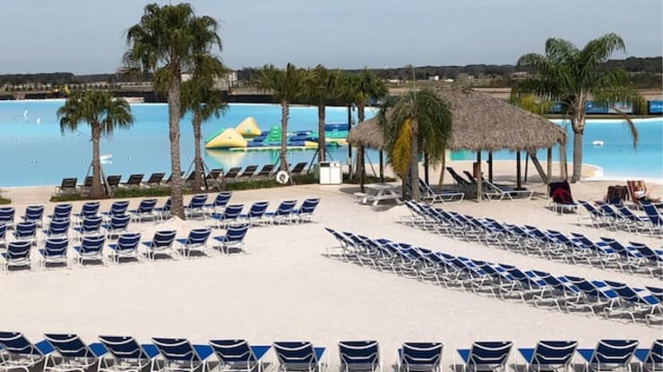 Beach resort with rows of blue lounge chairs, palm trees, and a thatched-roof shelter next to a large, blue lagoon.