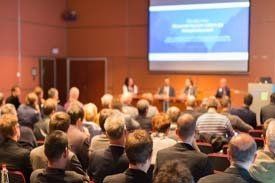 Audience in a conference hall, watching speakers at a table on a stage with a screen displaying information.
