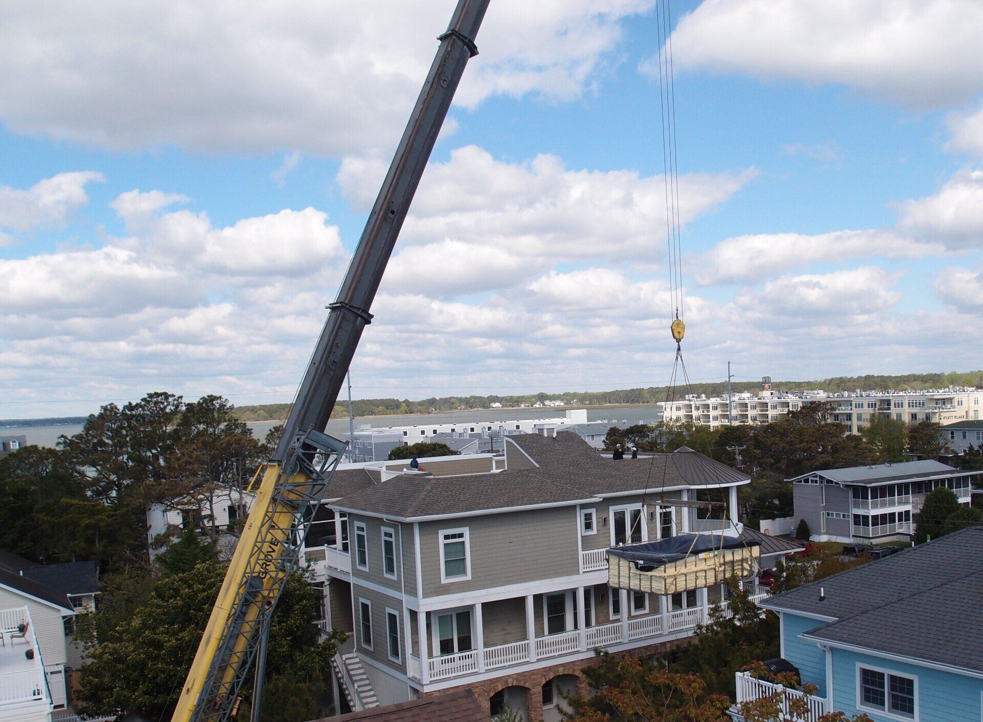 Crane lifting a house section; cloudy sky, houses and trees in the background.