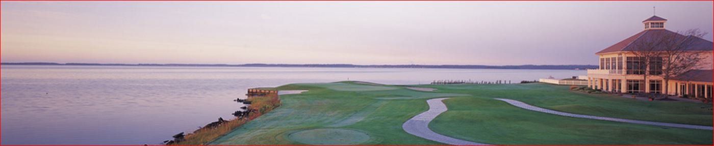 A building with a cupola overlooks a calm body of water, a walkway leading across the lawn.