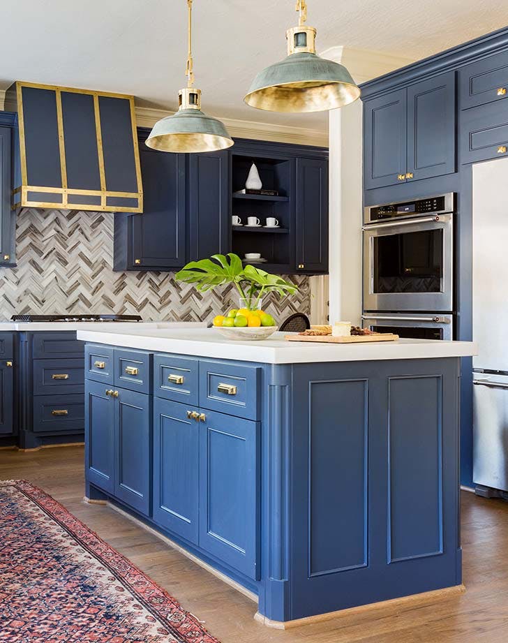 Blue kitchen with island, gold accents, herringbone backsplash, and hanging pendant lights.