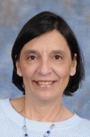 Woman with dark hair and light blue sweater smiles at the camera in front of a blurry blue background.