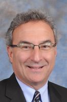 Smiling man with glasses, graying hair, wearing a dark suit and tie.