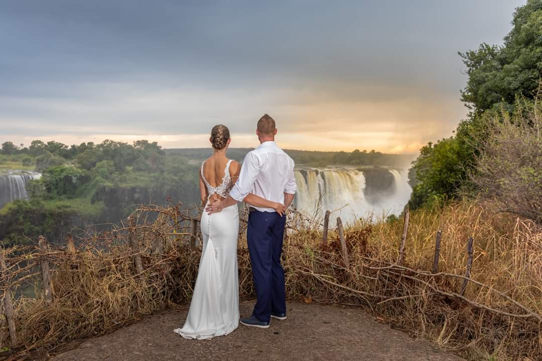 A bride and groom are standing in front of a waterfall.