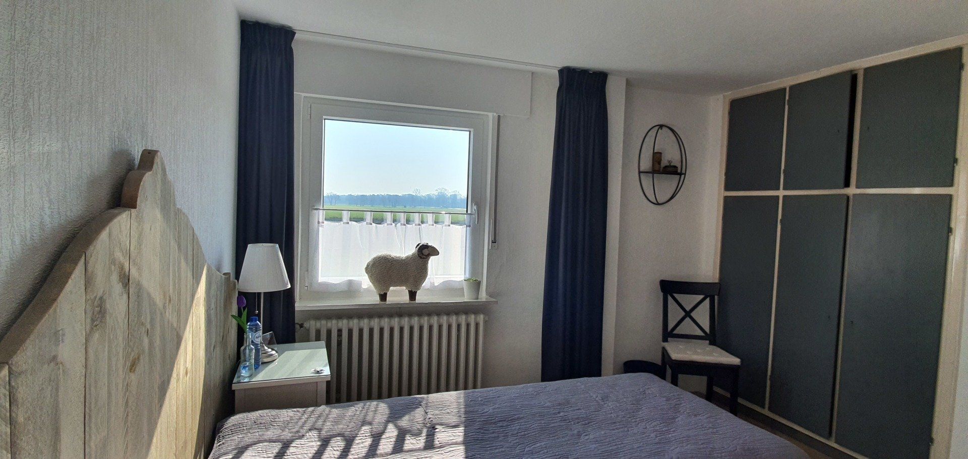 Slaapkamer met bed, kledingkast, gordijnen en een hond die op de vensterbank boven een radiator zit.