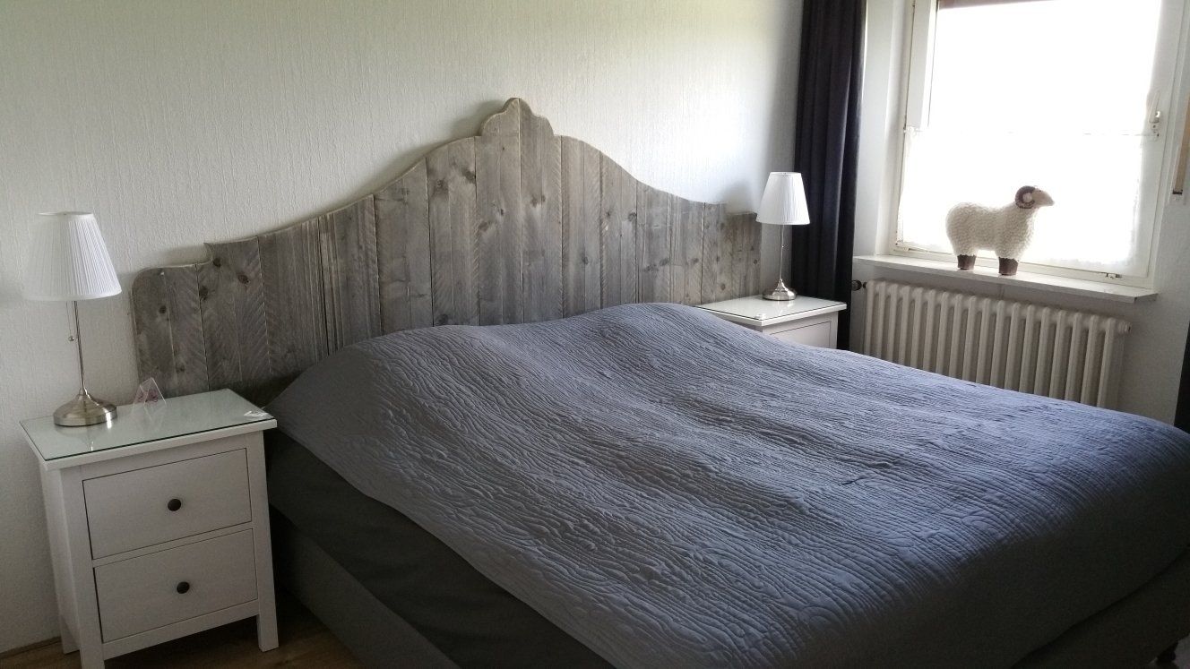 Slaapkamer met een grijze sprei, witte nachtkastjes en een klein hondje op de vensterbank.