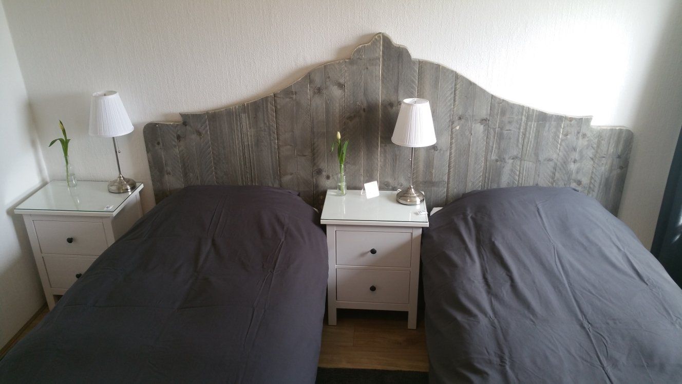 Twee keurig opgemaakte eenpersoonsbedden met donkergrijs beddengoed en een gedeeld houten hoofdeinde, geflankeerd door nachtkastjes en lampen.
