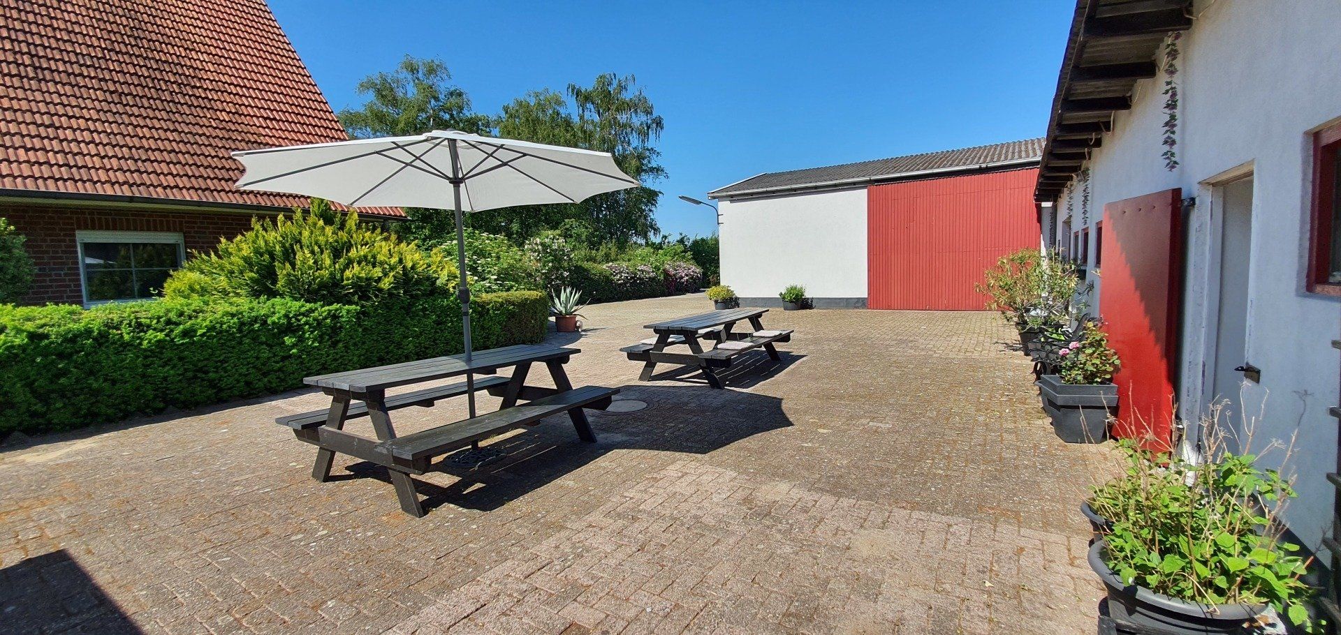 Zonnige binnenplaats met picknicktafels, witte parasols en rood-witte gebouwen.