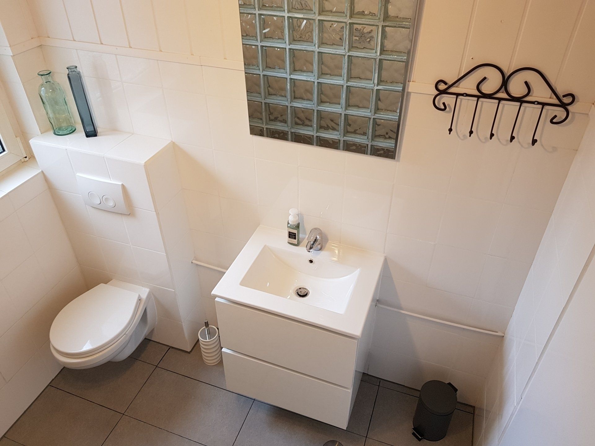 Kleine witte badkamer met toilet, wastafelmeubel, raam van glazen bouwstenen en handdoekhaak aan betegelde muren.