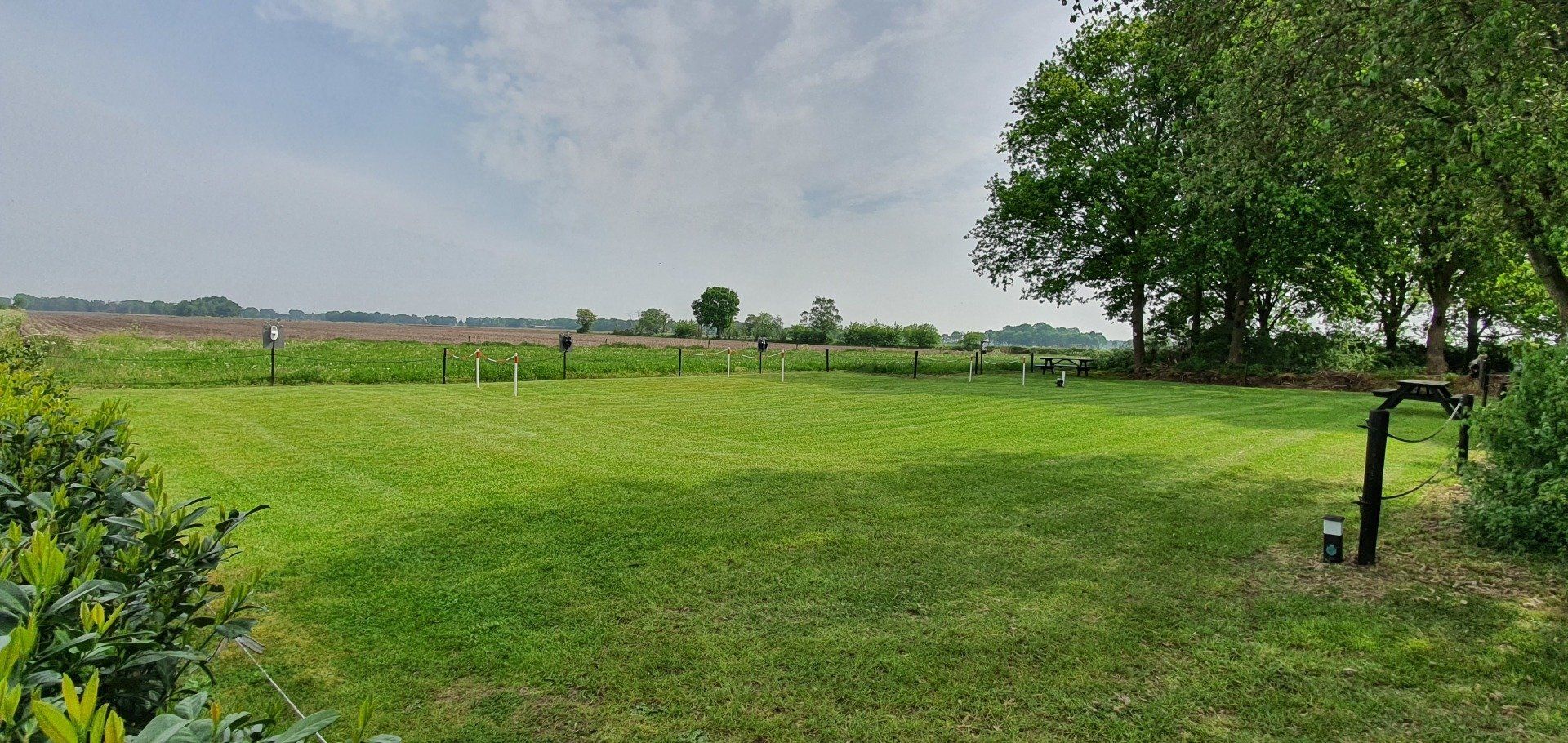 Een uitgestrekt groen grasveld onder een bewolkte hemel, met bomen en een hek in de verte.