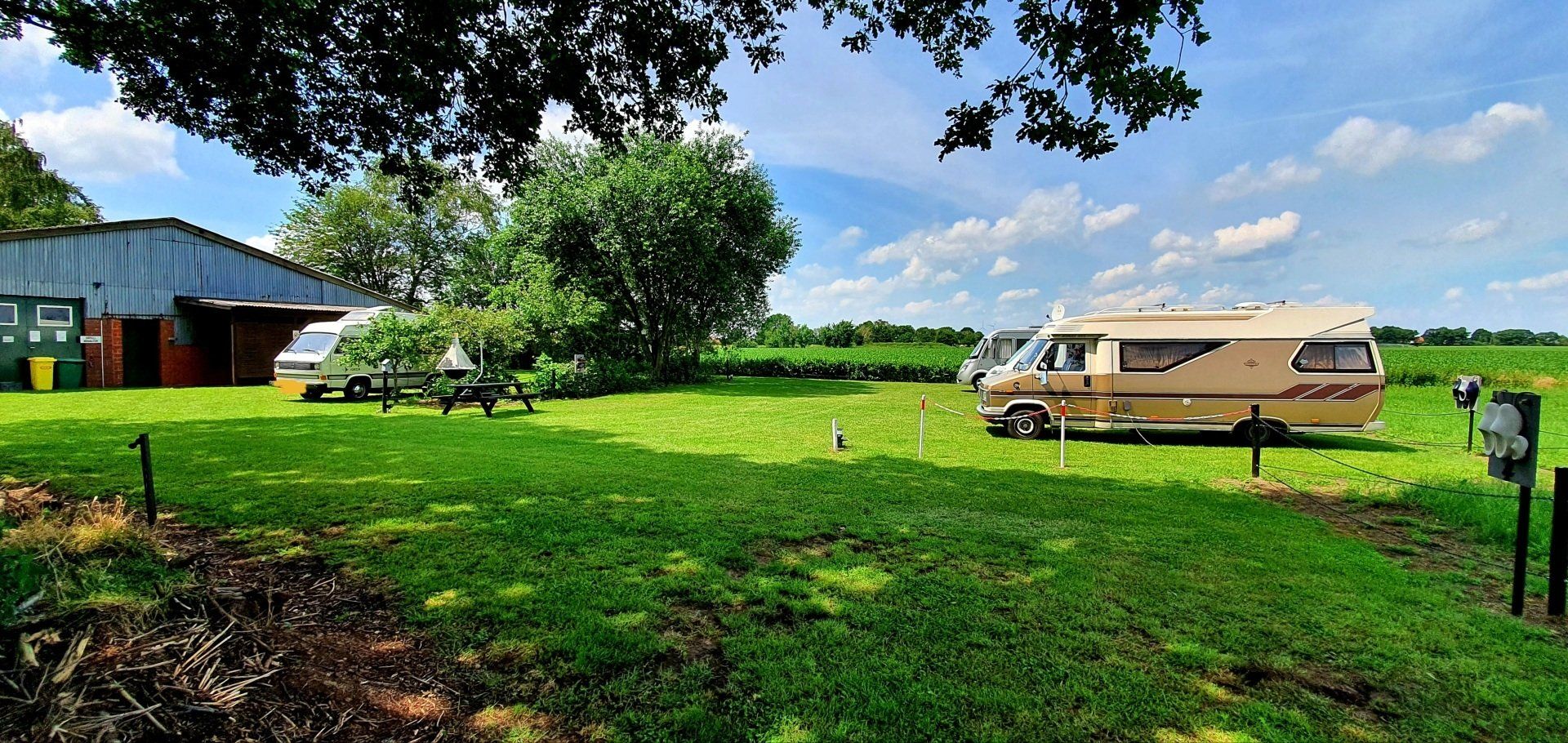 Een groen landelijk veld met een beige bestelwagen, een schuur en bomen onder een bewolkte hemel.