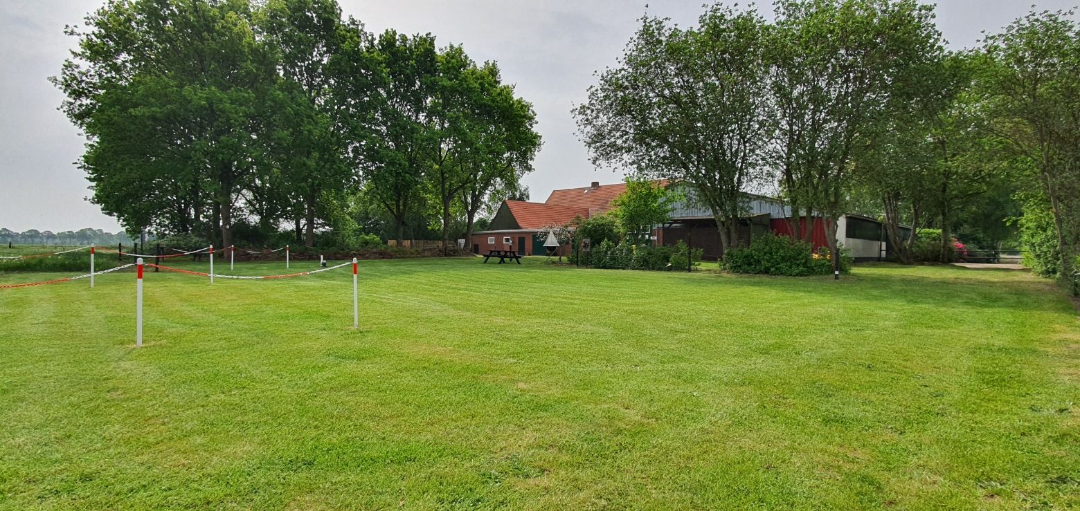 Groen grasveld met witte palen, bomen en een huis op de achtergrond onder een bewolkte hemel.