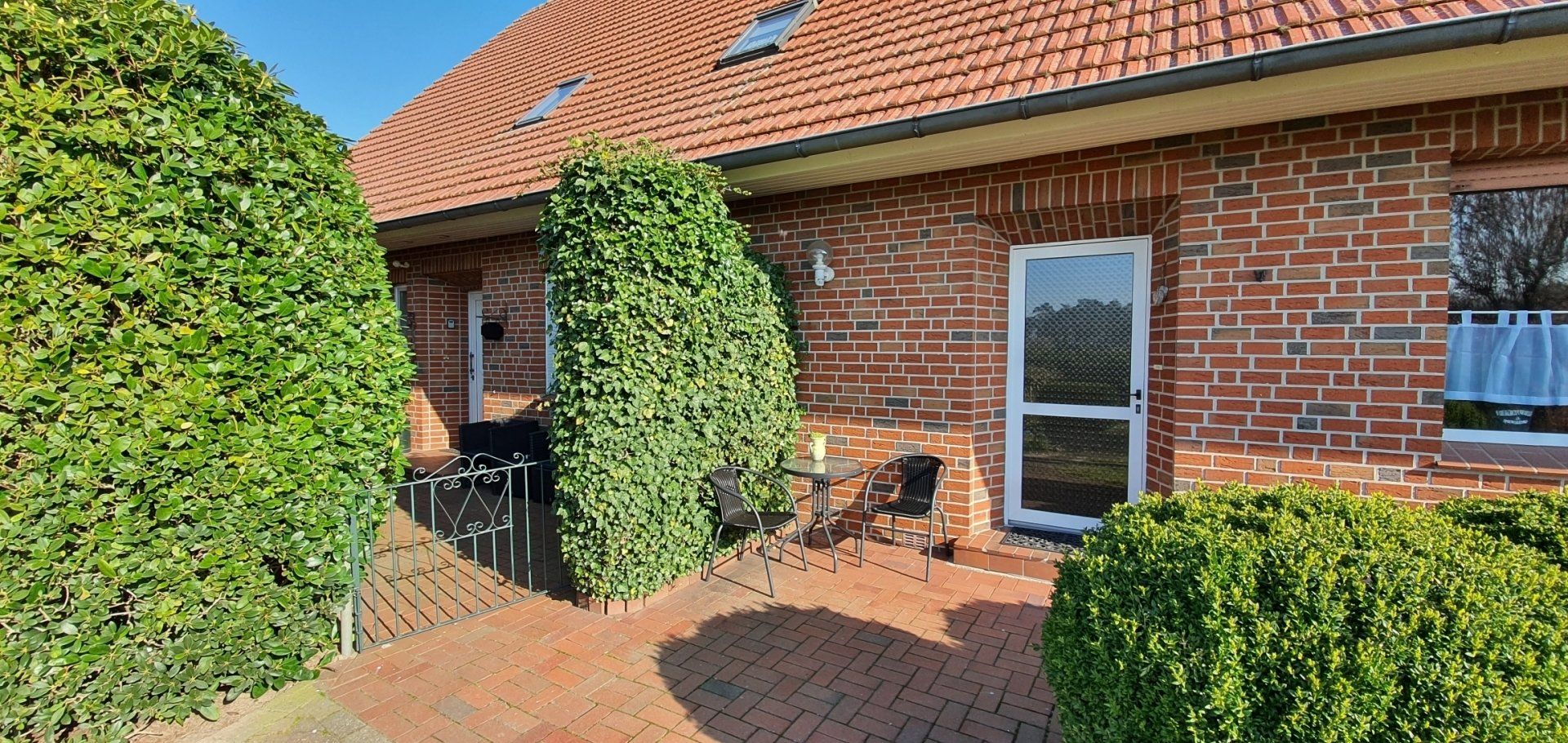 Bakstenen huis met rood pannendak, groene hagen en een betegeld terras aan de voorzijde in de volle zon.