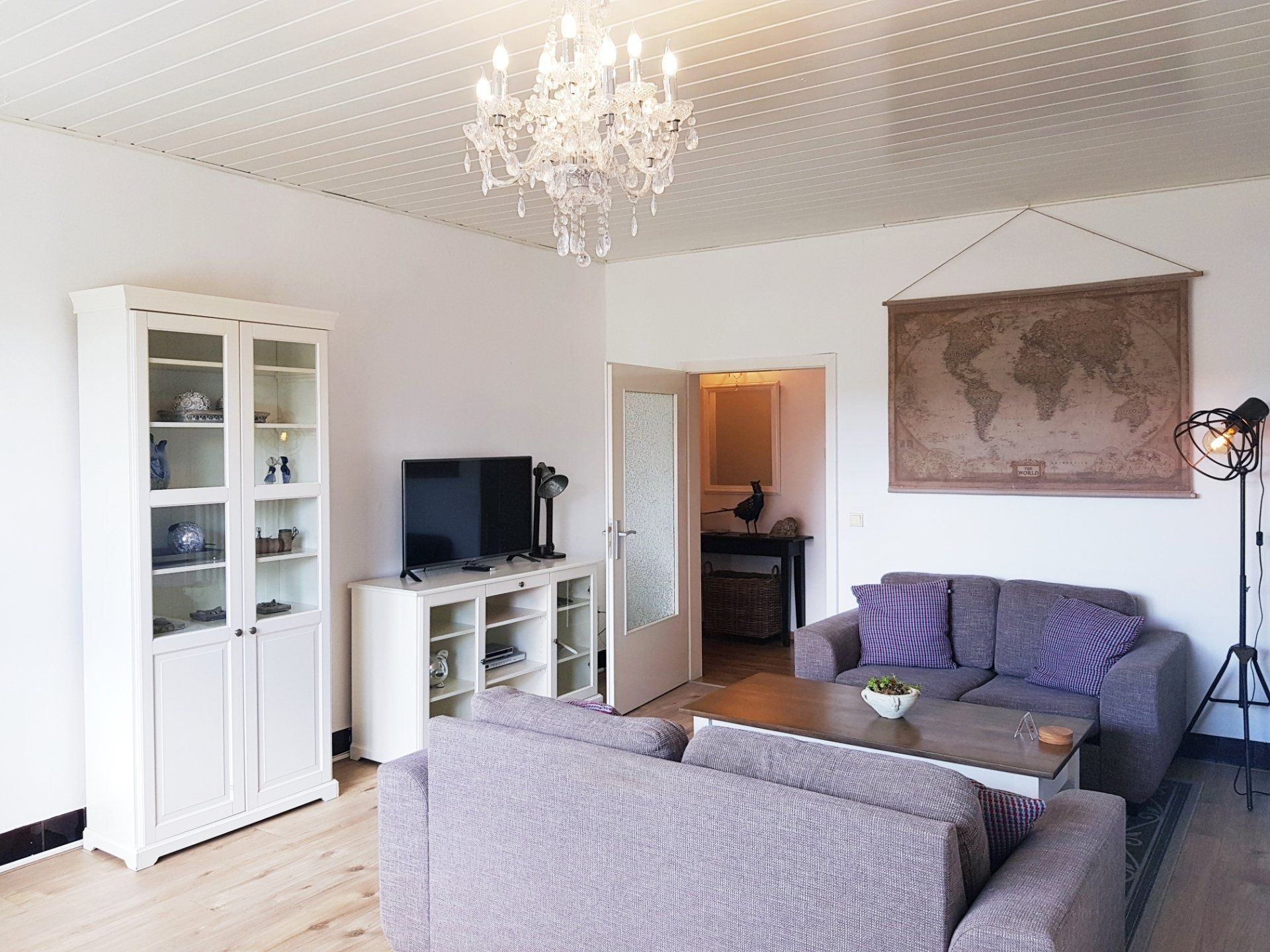 Lichte woonkamer met grijze bank, witte kast, tv, kroonluchter en een wereldkaart aan de muur.