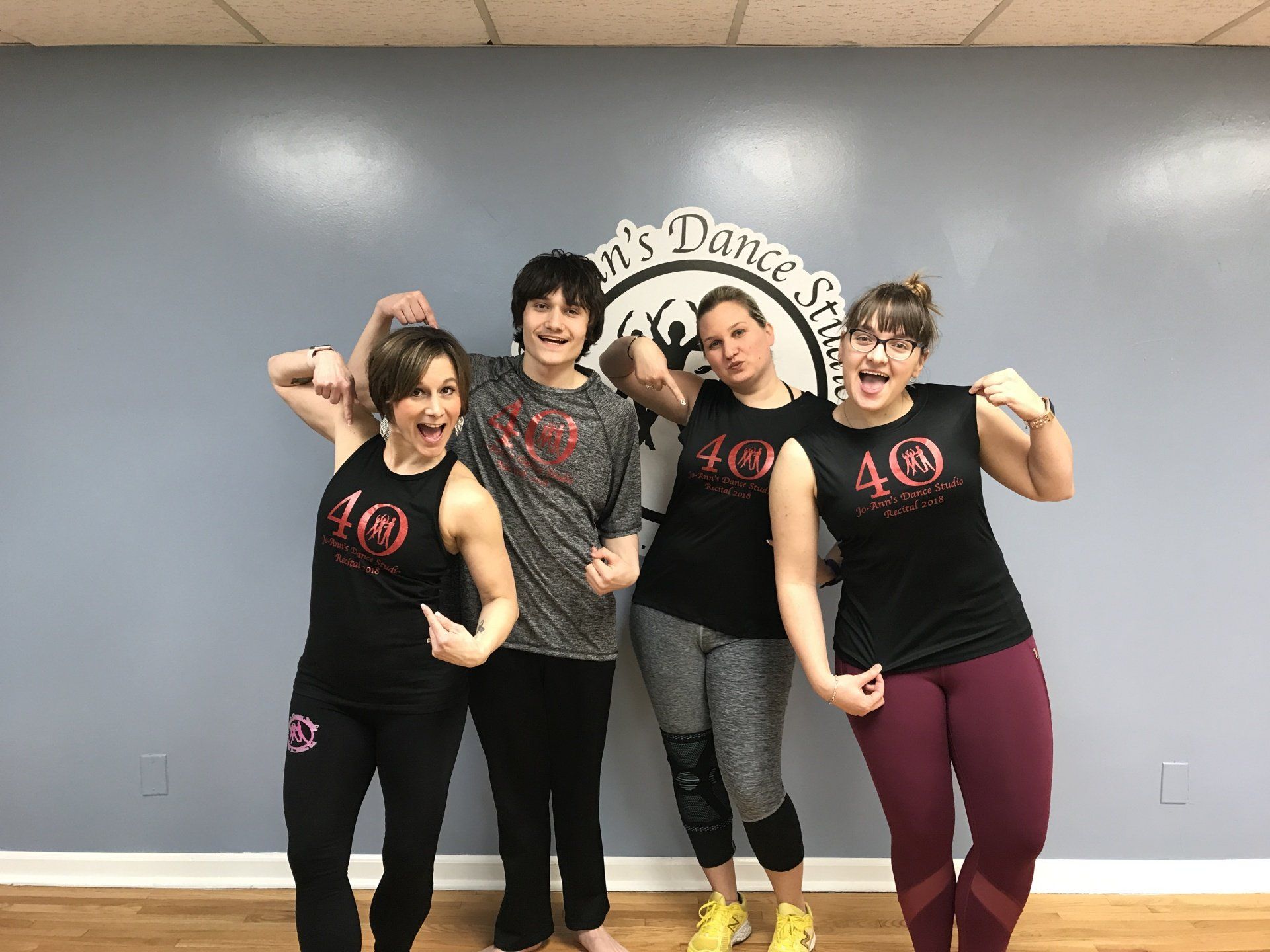 Four dancers in black and maroon outfits pose flexing against a gray studio wall.