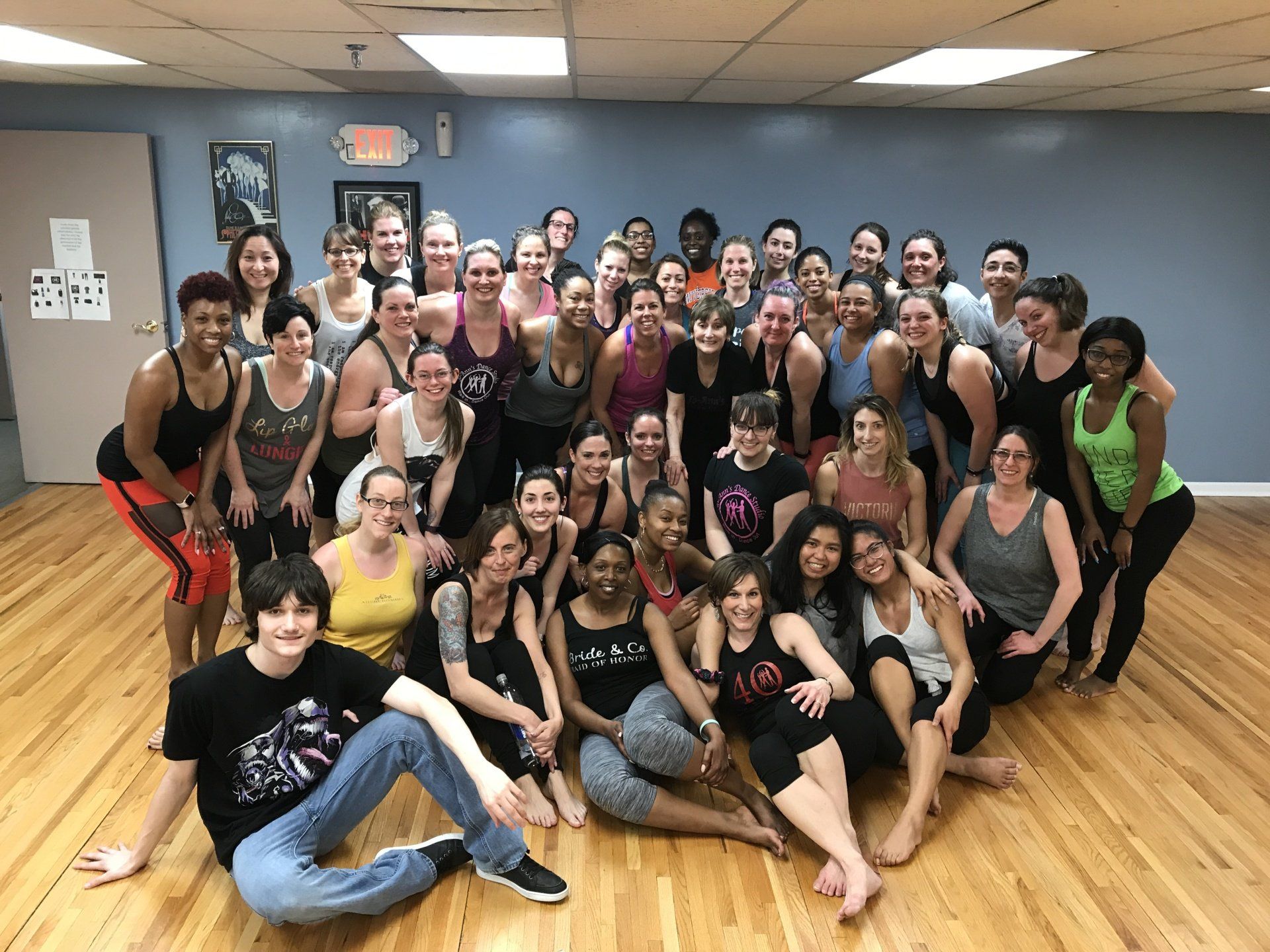Large group posing in a dance studio, many smiling and seated on a wooden floor with mirrors behind them