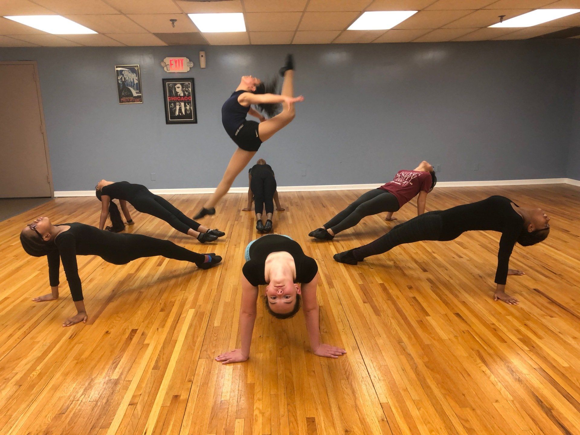 Five dancers in black and maroon hold acrobatic poses on a wooden studio floor, one centered in a high kick.