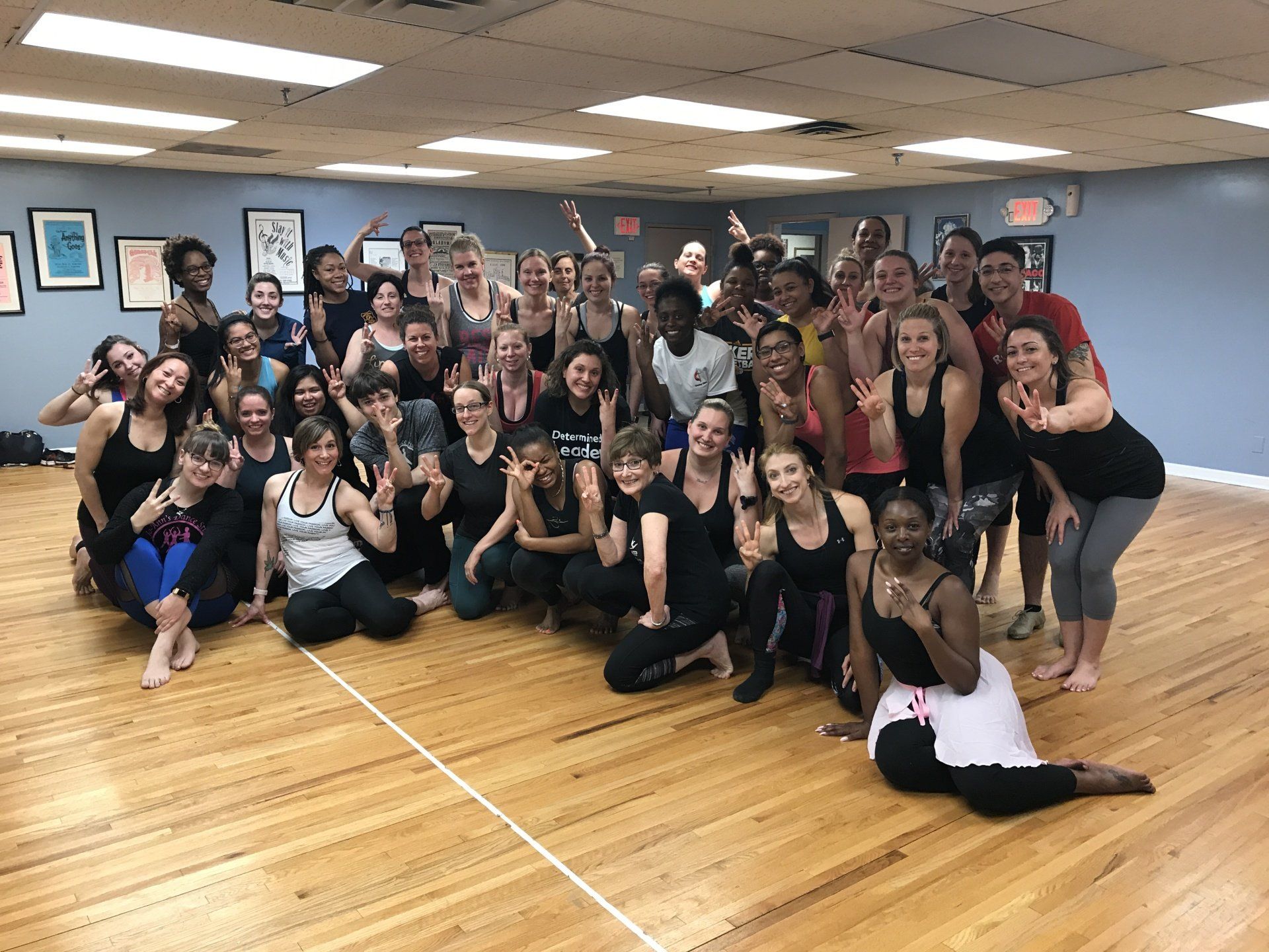 Large group posing in a dance studio, many smiling and crouching on a wooden floor with mirrors and posters behind them